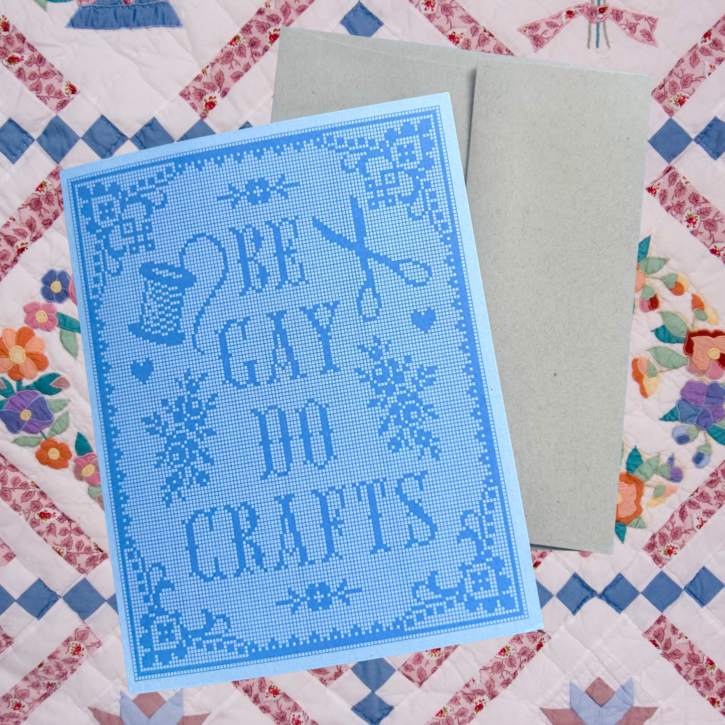 Blue cross-stitched card with 'Be Gay Do Crafts' text on a Aida fabric background